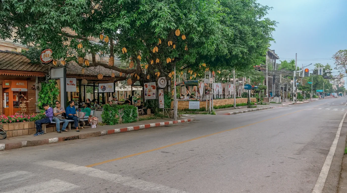 Chiang Mai street with cafes and community life Street view of Chiang Mai with cafes, shops, and people sitting under a tree, showcasing the local community atmosphere